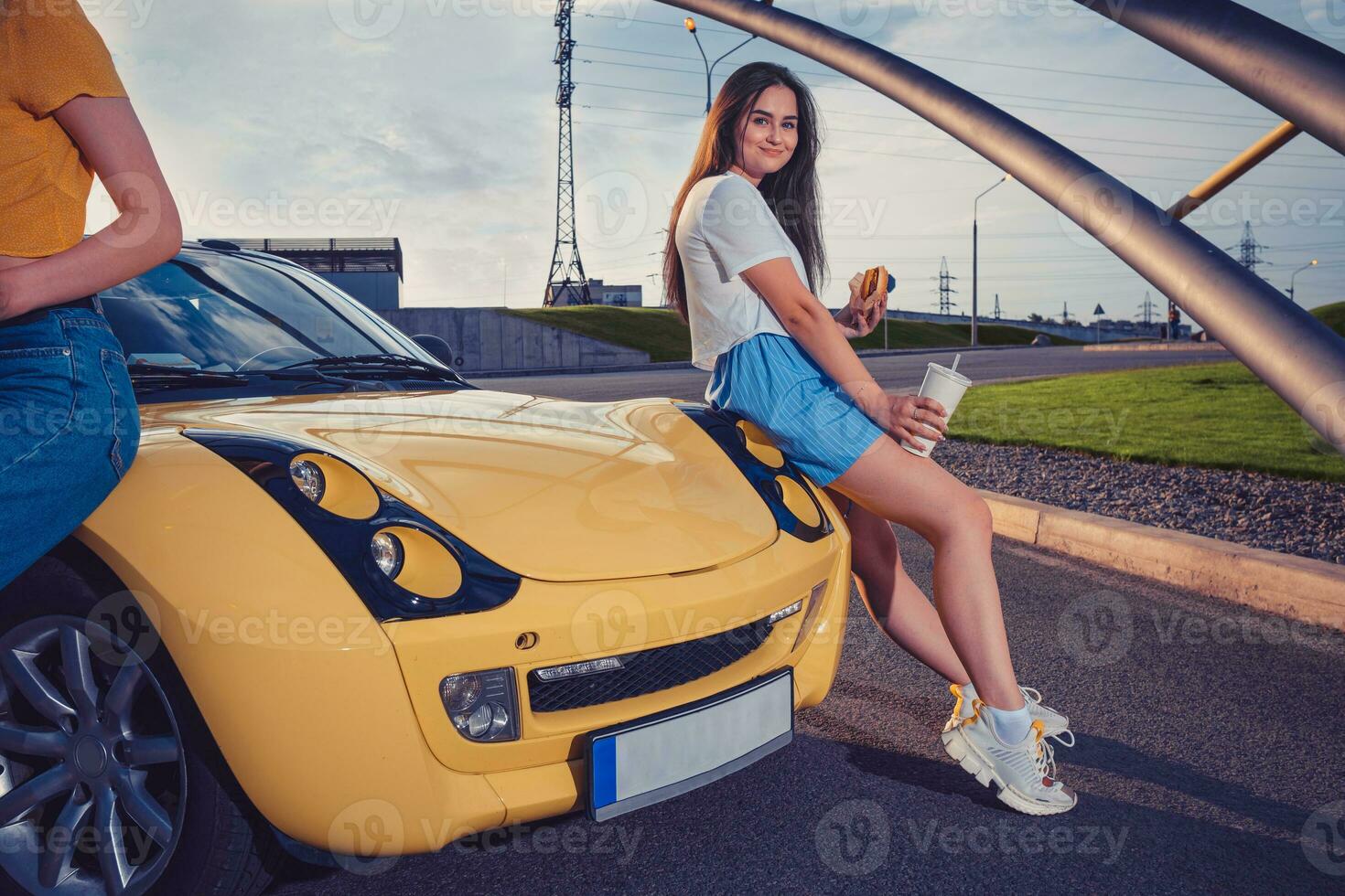 Female in casual outfit is holding hamburger and drink in paper cup, sitting on hood of yellow car with her friend. Fast food. Close up, copy space photo