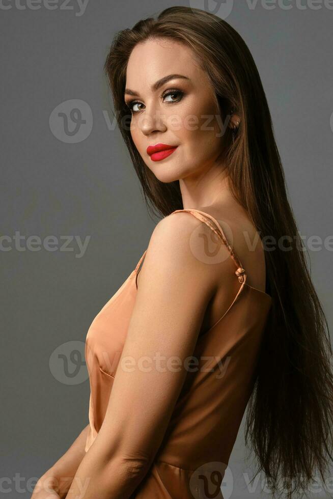 Brunette model with bright make-up and long hair, in silk golden dress. Smiling while posing sideways against gray background. Close up photo