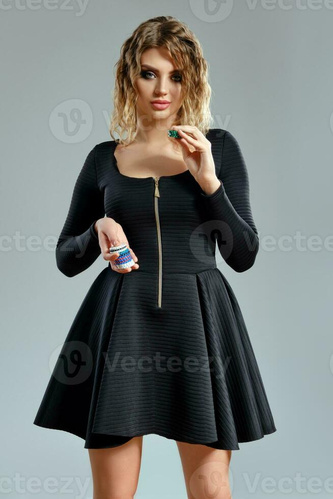 Blonde lady in black short dress showing some colored chips, posing against gray background. Gambling entertainment, poker, casino. Close-up. photo