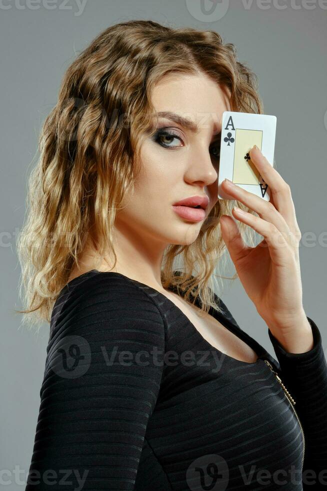 Maiden in black dress has covered her eye with ace of clubs while posing sideways on gray background. Gambling entertainment, poker, casino. Close-up. photo
