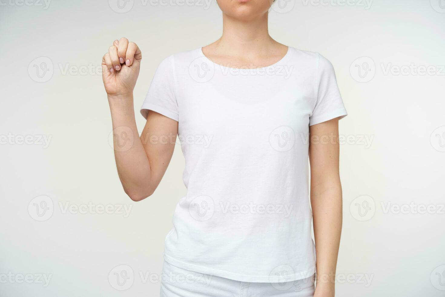 Horizontal shot of young lady dressed in casual clothes keeping hand raised while showing letter n using deaf alphabet, standing over white background photo