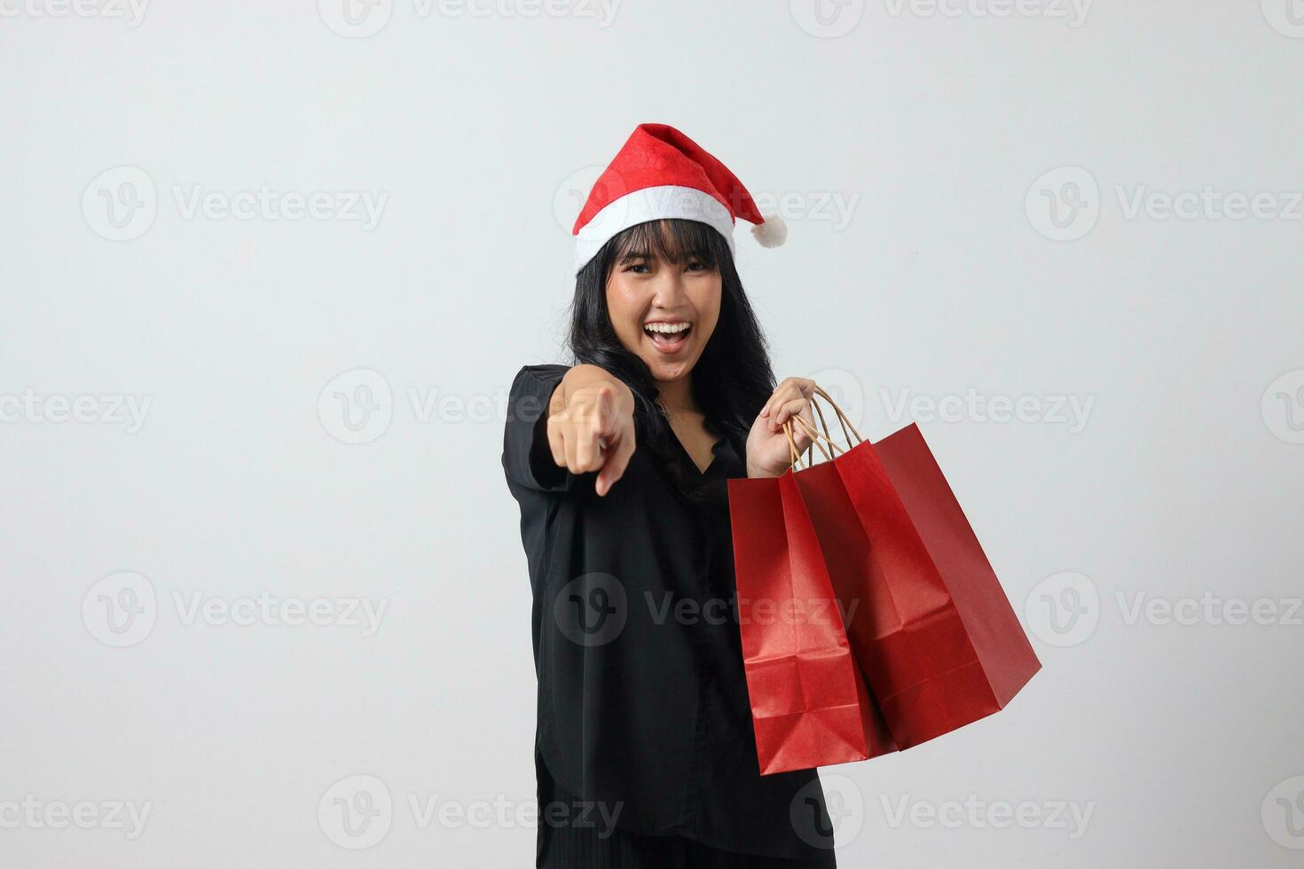 retrato de alegre asiático mujer con rojo Papa Noel sombrero sensación contento mientras señalando a el lado. emocionado niña participación compras bolsa. nuevo año y Navidad concepto. aislado imagen en blanco antecedentes foto