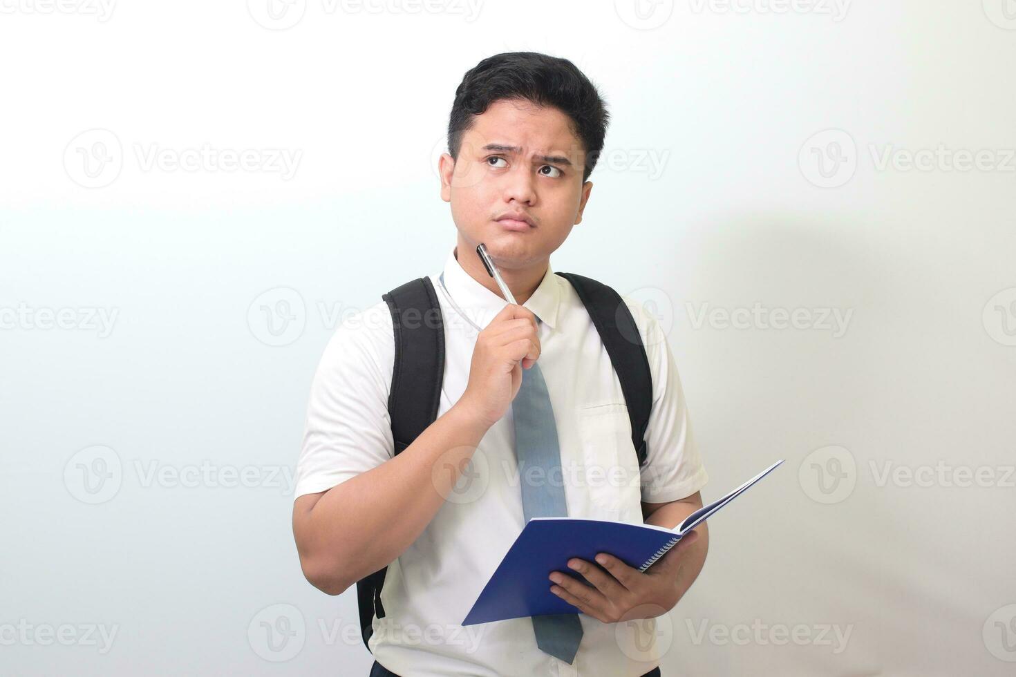 Indonesian senior high school student wearing white shirt uniform with gray tie writing on note book using pen and thinking about an idea. Isolated image on white background photo