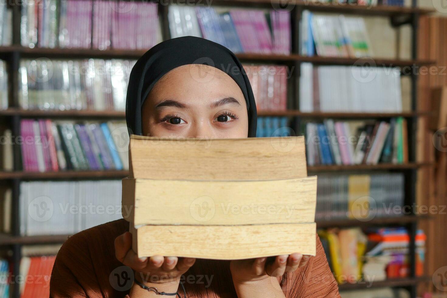 Portrait of Asian hijab woman covering her head with a pile of books in front of library bookshelf. Muslim girl reading a book. Concept of literacy and knowledge photo