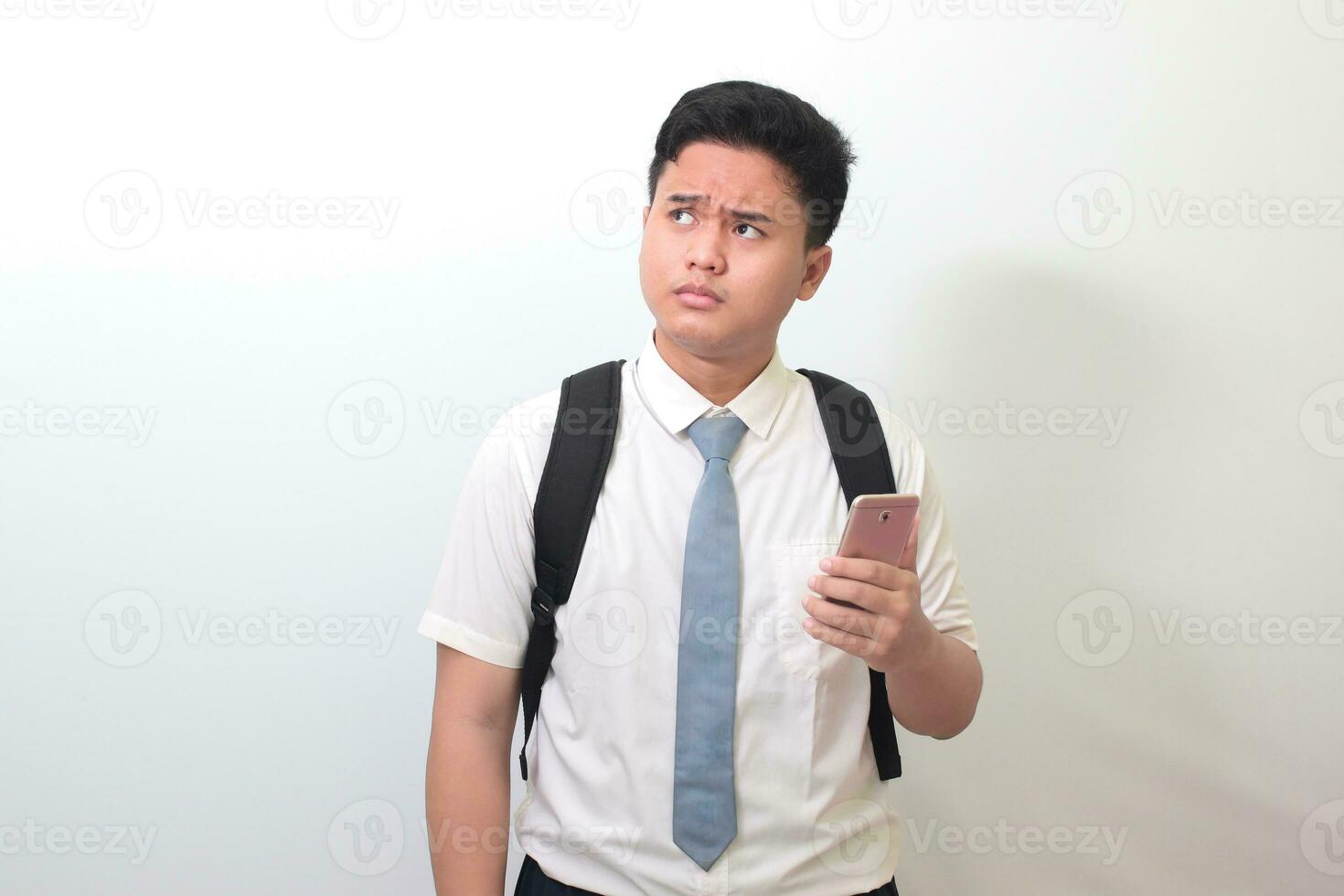 Indonesian senior high school student wearing white shirt uniform with gray tie showing confusion face expression while holding a mobile phone. Isolated image on white background photo