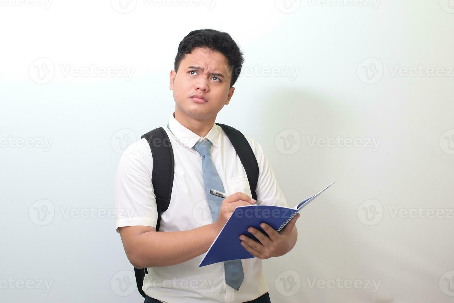 Indonesian senior high school student wearing white shirt uniform with gray tie writing on note book using pen and thinking about an idea. Isolated image on white background photo