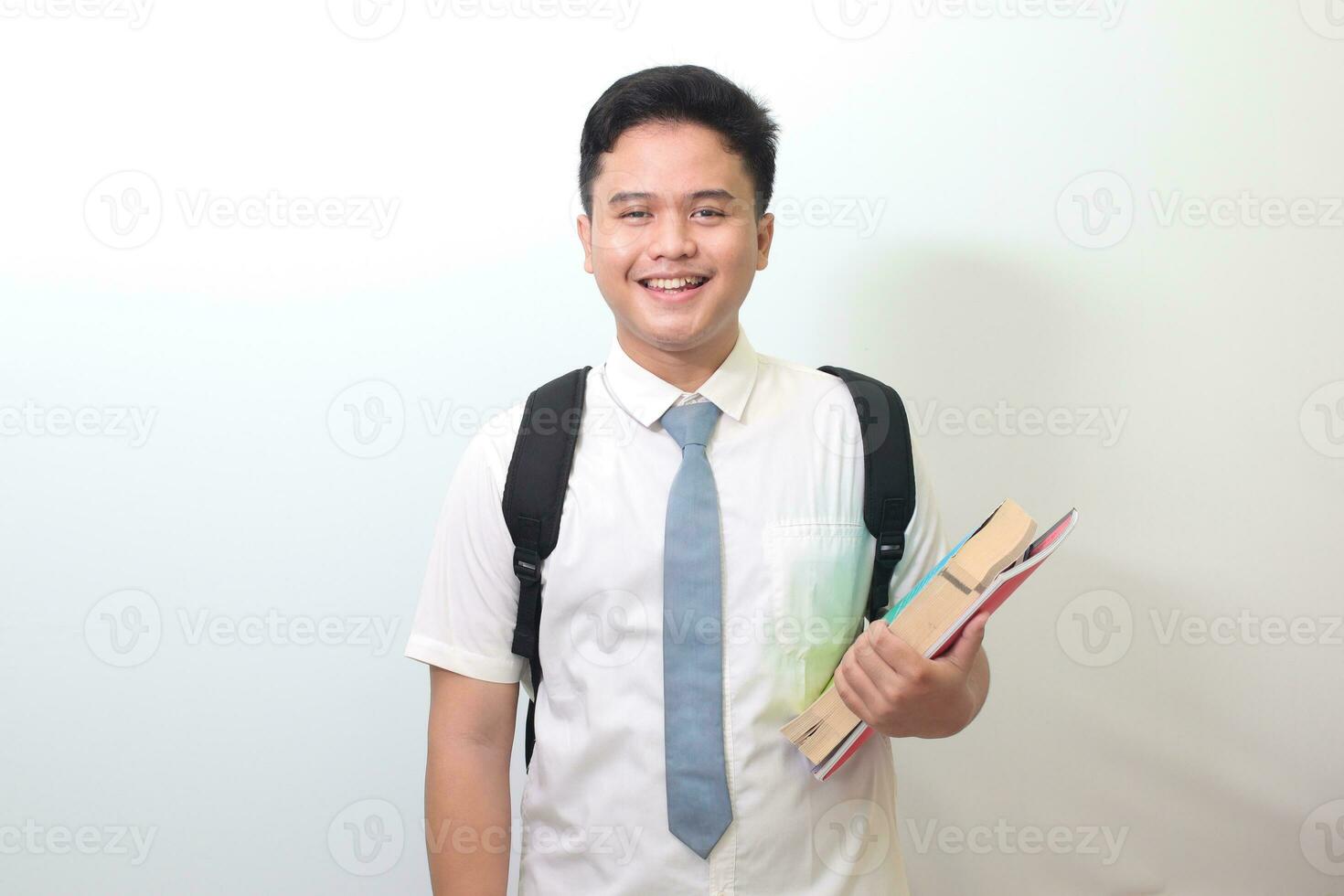 indonesio mayor alto colegio estudiante vistiendo blanco camisa uniforme con gris Corbata participación algunos libros, sonriente y mirando a cámara. aislado imagen en blanco antecedentes foto