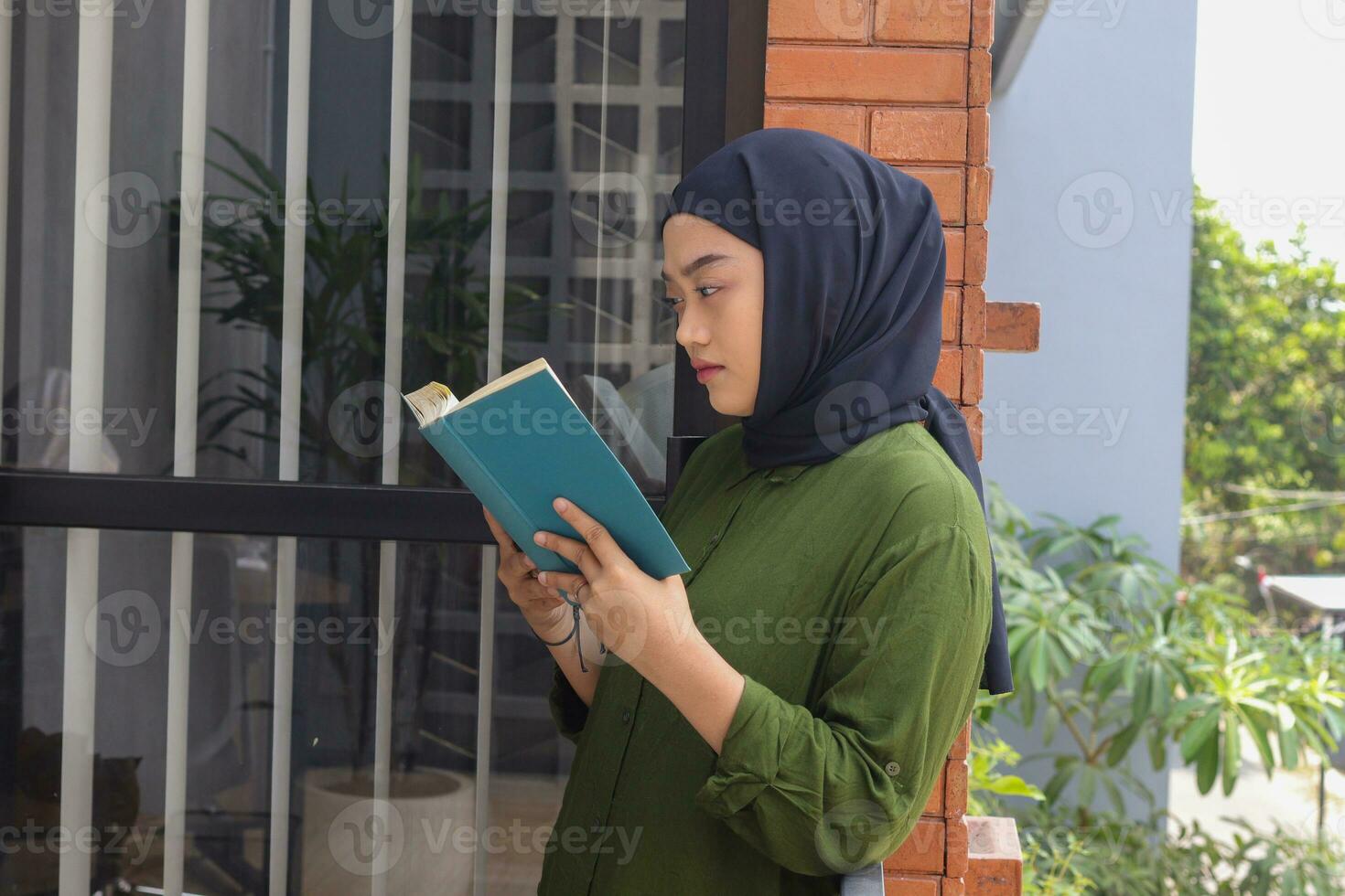 Portrait of attractive Asian hijab woman holding book in balcony. Muslim girl reading a book. Concept of literacy and knowledge photo