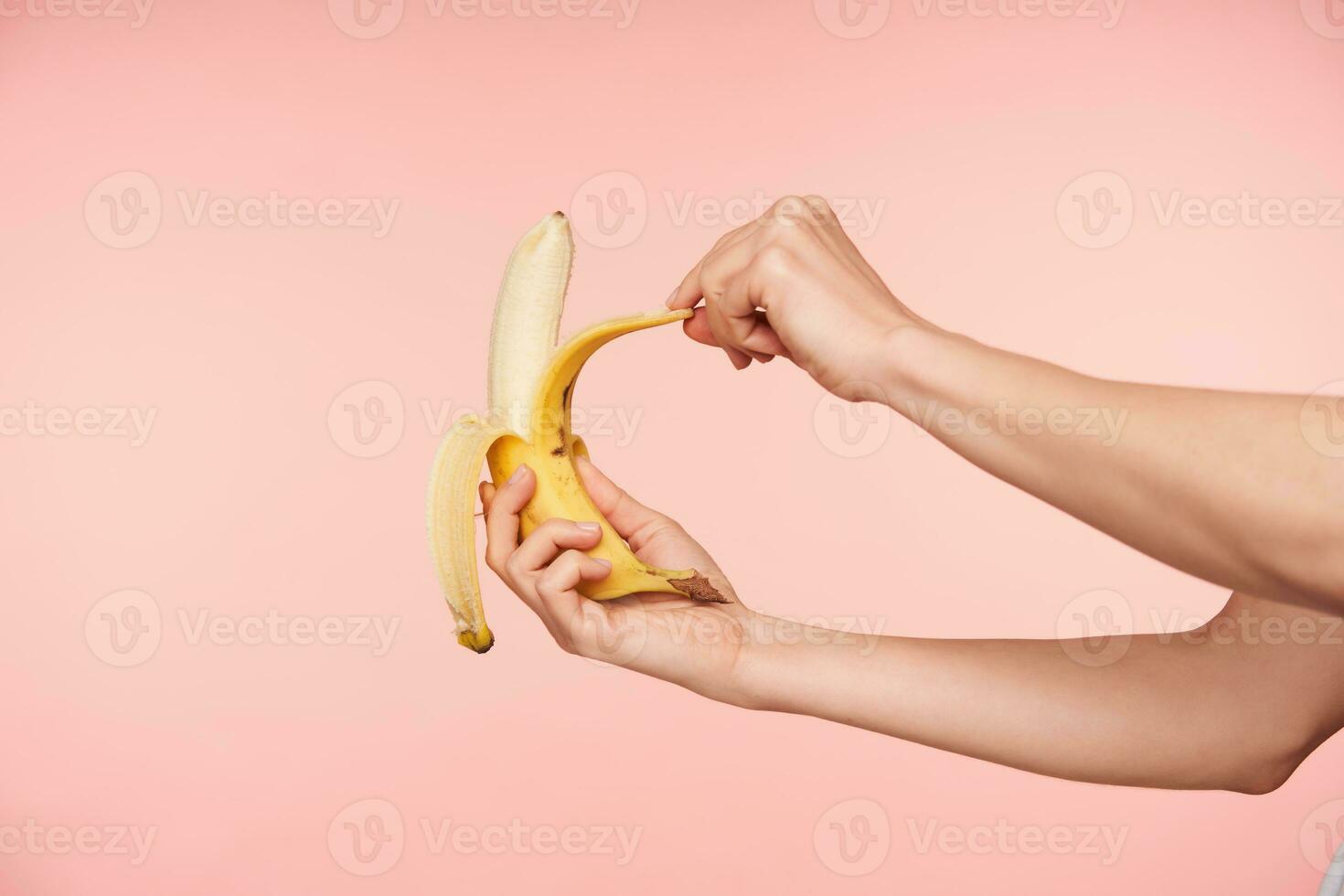 Studio shot of elegant woman's hands holding banana while peeling it and going to bite, having healthy breakfast while being isolated over pink background photo