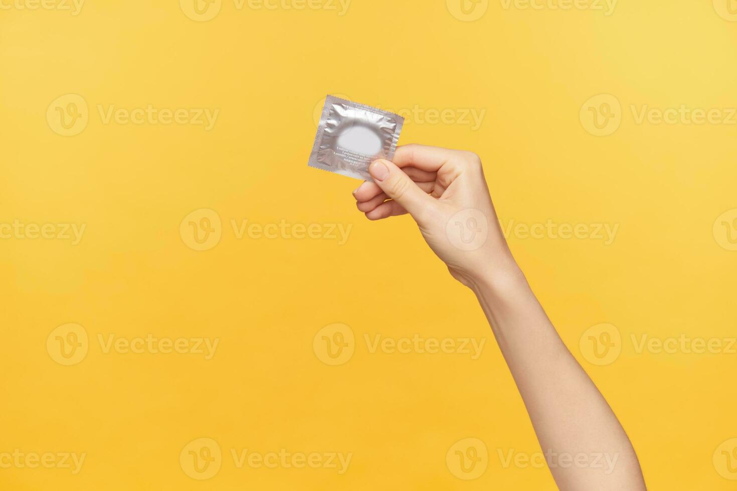 Horizontal shot of young fair-skinned female's hand being raised while holding silver pack with condom. Young woman prefer safe sex, posing over orange background photo