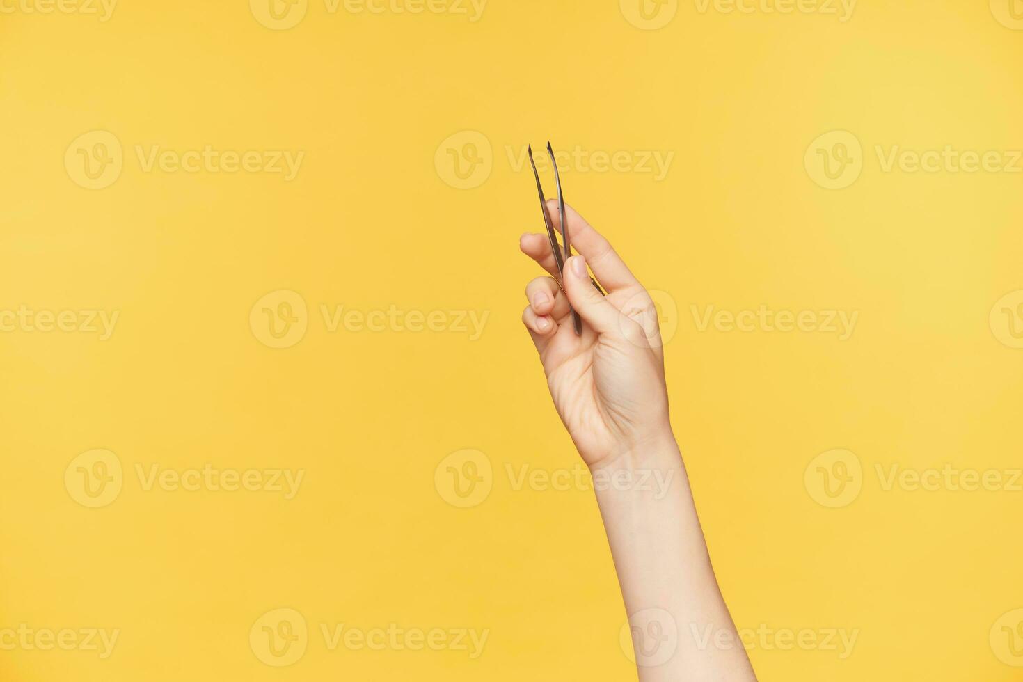 Studio shot of young female's well-groomed hands keeping tweezers while posing over orange background. Young woman is going to form her eyebrows photo