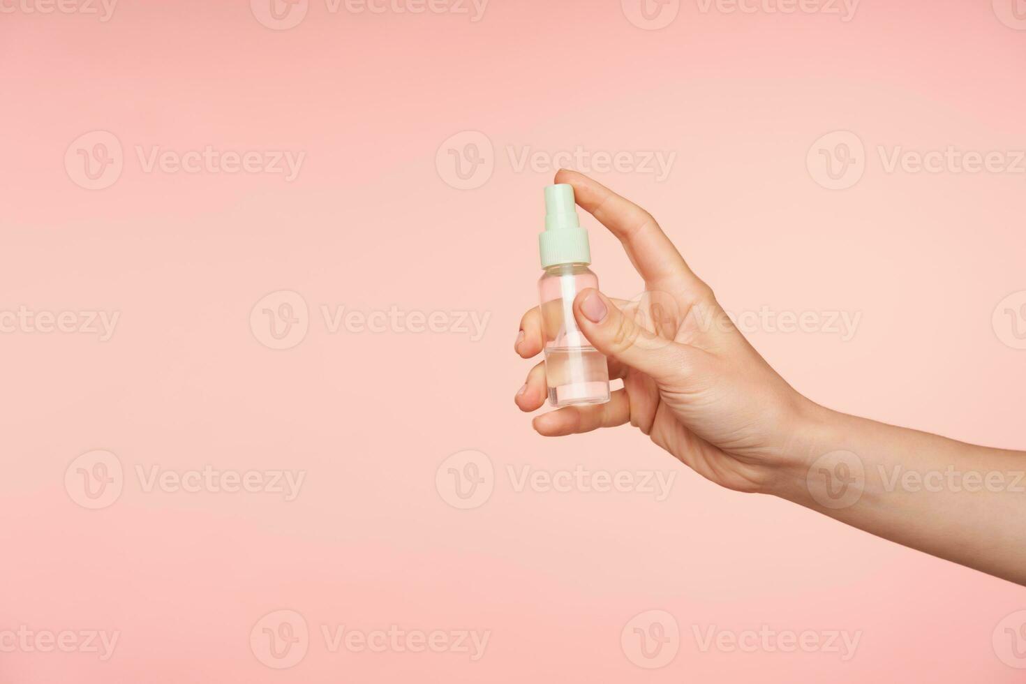 estudio foto de joven femenino Piel blanca mano acuerdo dedo índice en botón de botella mientras yendo a rociar contenido, aislado terminado rosado antecedentes