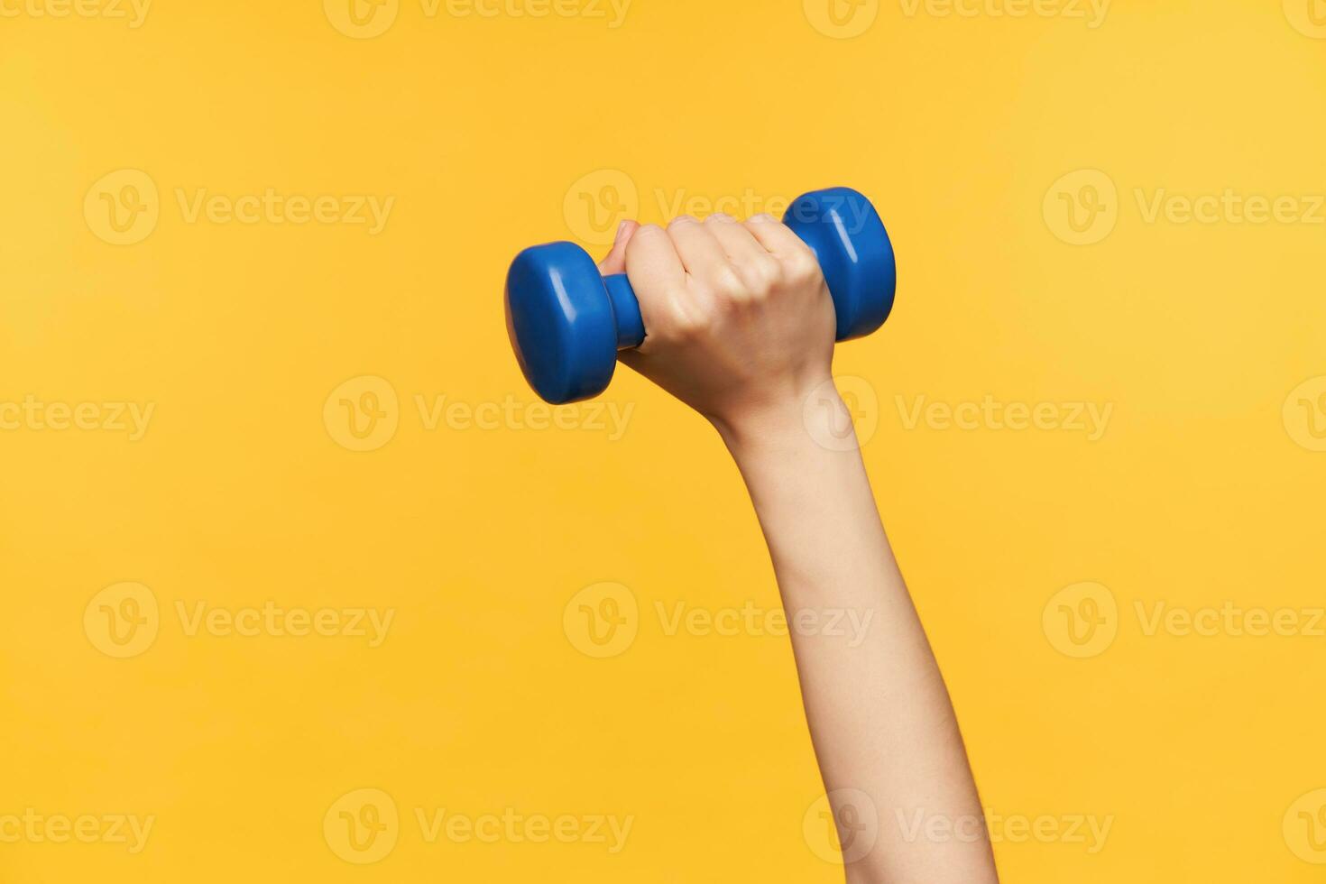 Studip photo of blue dumbbell being kept by fair-skinned woman hand while making workout for arms, isolated over yellow background. Body care and sports concept