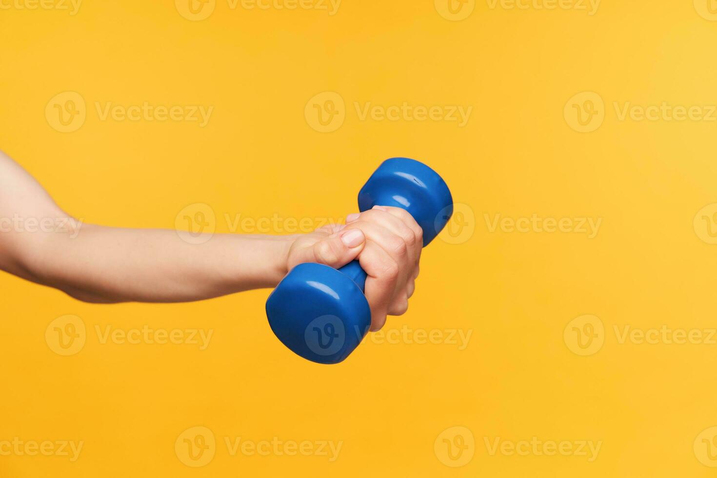 Side view of young fair-skinned woman hand with nude manicure keeping blue dumbbell in it while having fitness class, isolated over yellow background photo