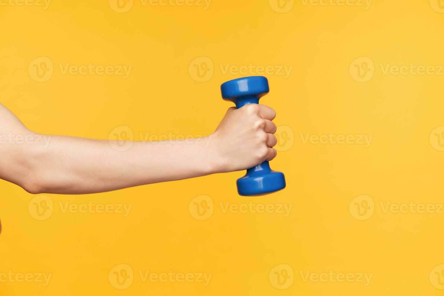 Side view of raised female hand with blue dumbbell exercising biceps while being isolated over yellow background. Body care and fitness concept photo