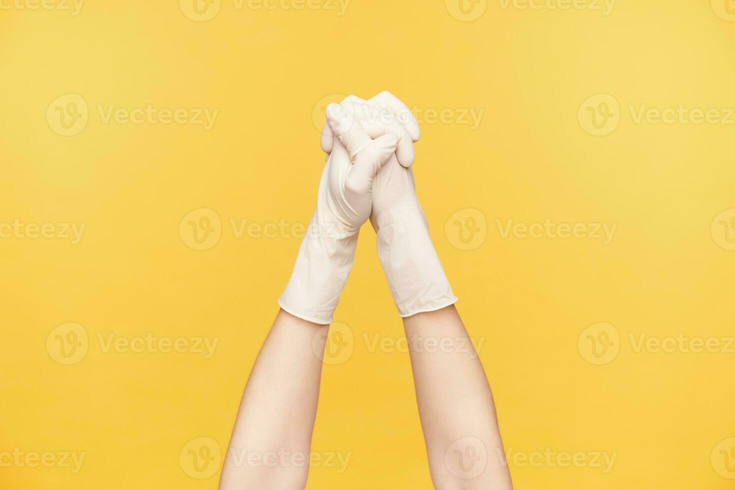 Studio shot of raised young woman's hands in rubber gloves folding together and keeping fingers crossed while being isolated over orange background photo