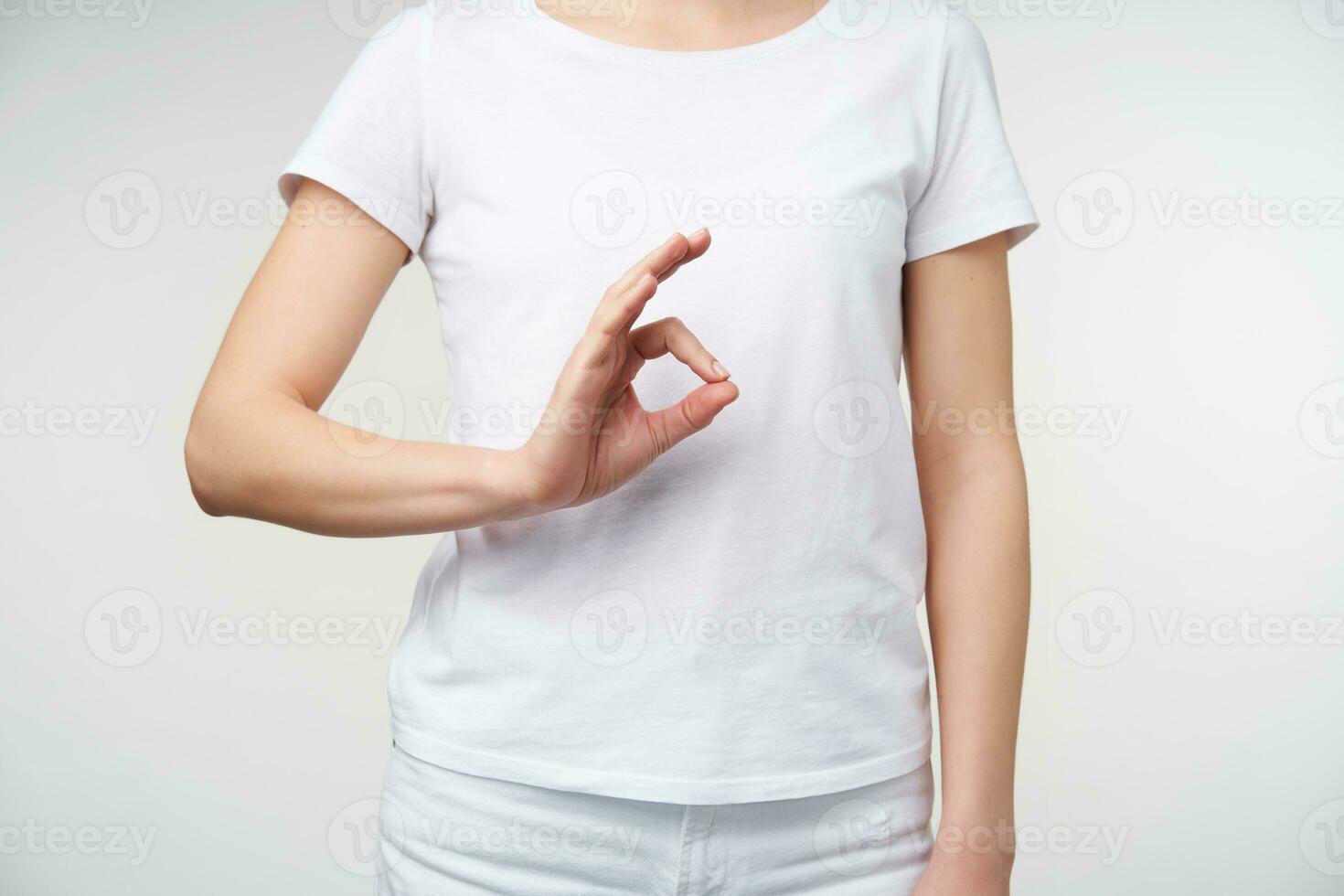 Indoor photo of young woman dressed in casual clothes forming ok gesture with her fingers while standing over white background. Sign language concept