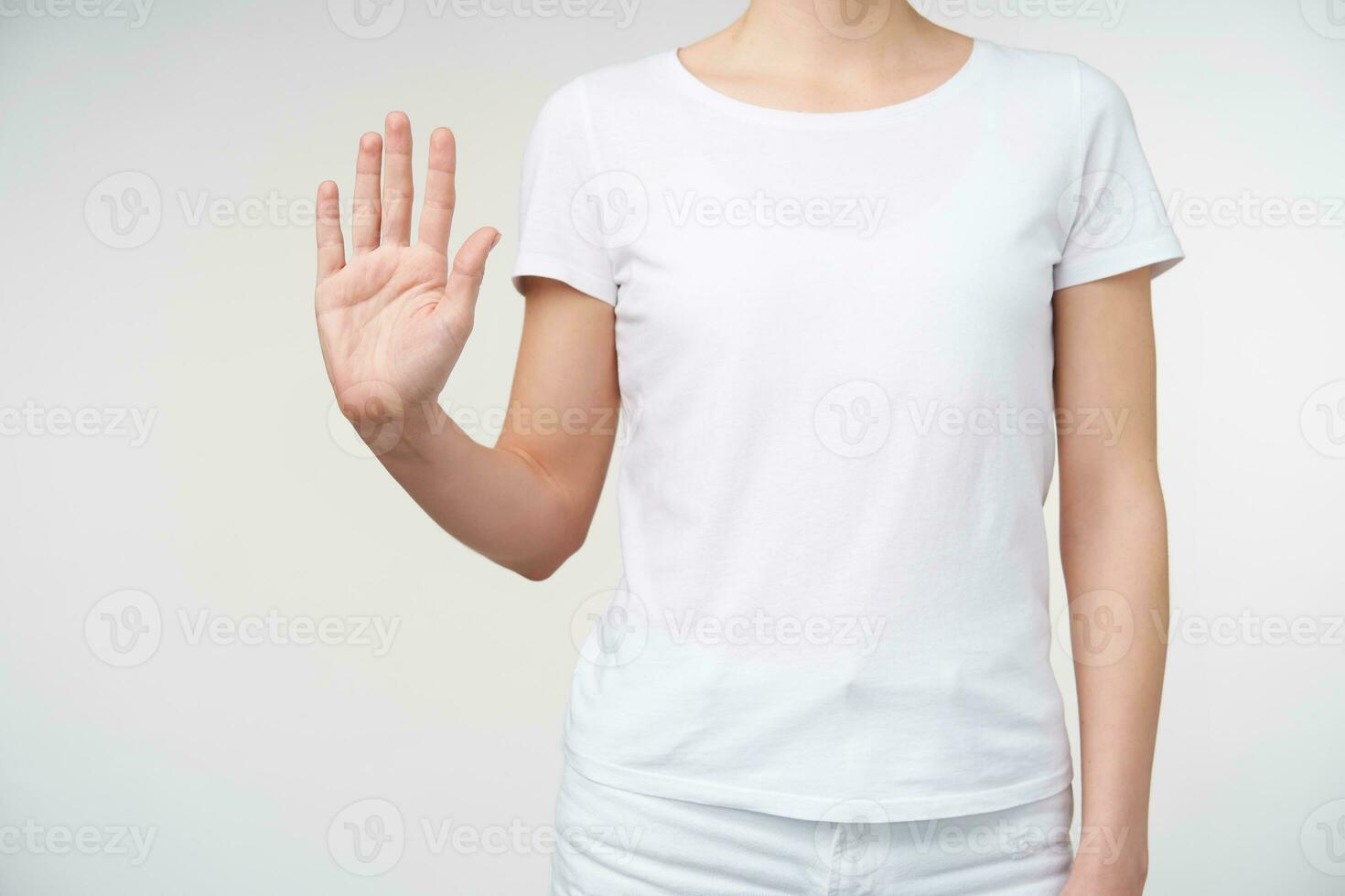 Shot of young female dressed in white t-shirt showing raised palm at camera while pointing up with fingers number five, standing over white background photo