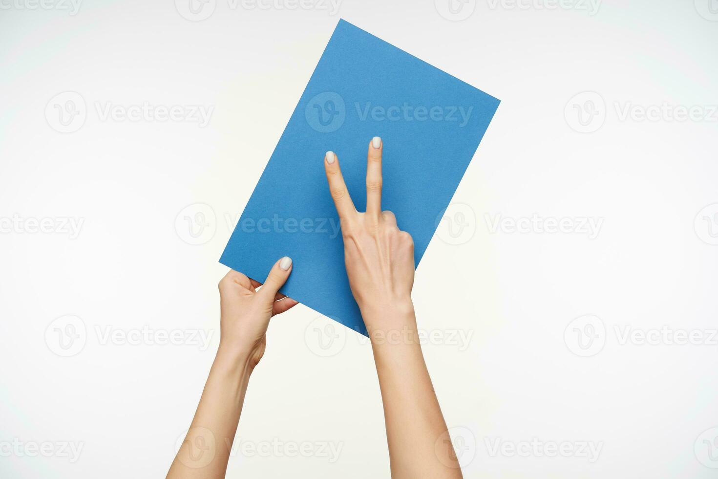 Front view of elegant young female's hands forming victory gesture with raised fingers and keeping piece of blue paper while standing over white background photo