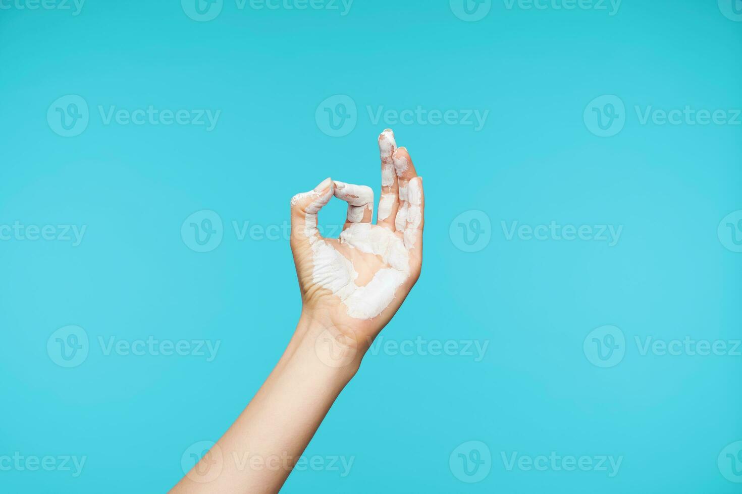 Photo of painted in white color hand being raised while showing mudra gesture, posing over blue background while meditating. Body language concept