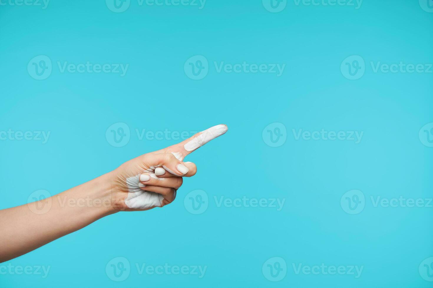 Close-up of young pretty lady's hand painted with white color keeping index finger raised while showing aside, posing against blue background photo