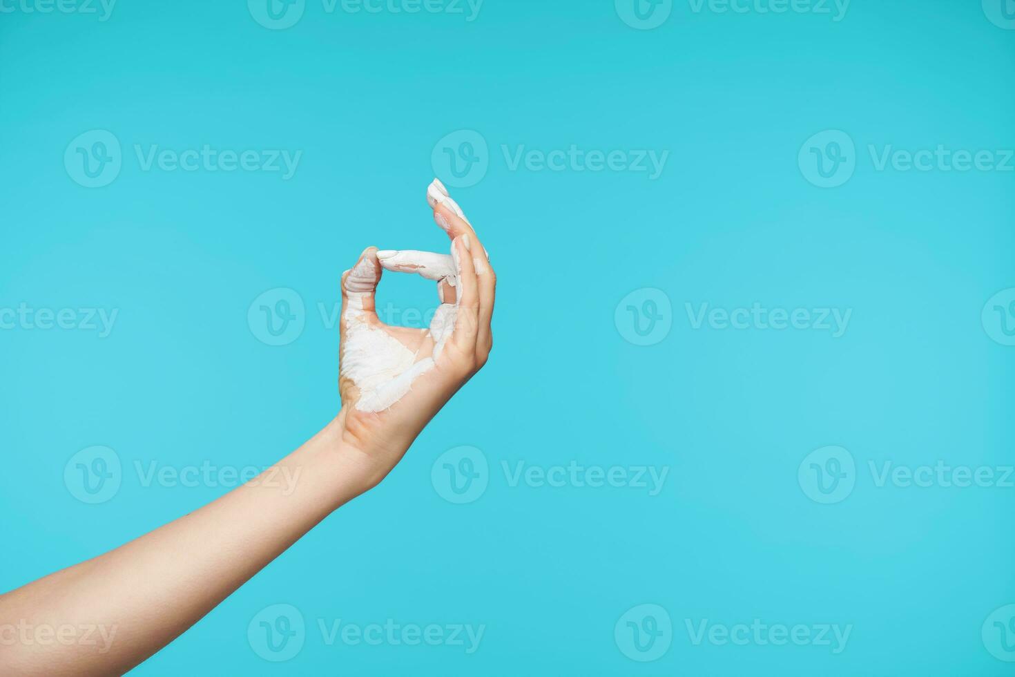 Studio photo of raised hand colored in white forming fingers in namaste sign while practicing yoga on early morning, posing over blue background