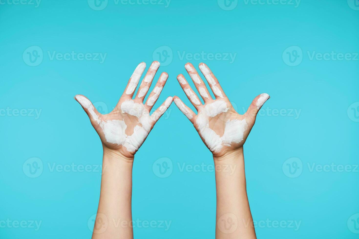 Two young female's hands showing up palms with white paint on it and keeping all fingers separately, isolated against blue background. Human gestures concept photo