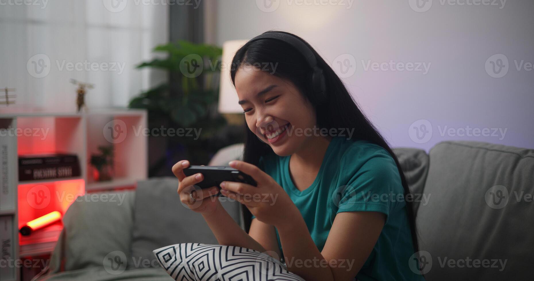 Portrait of Young Asian woman wearing glasses and headphones enjoys playing online esport games on smartphone sitting on sofa in the living room at home,Gamer lifestyle concept. photo