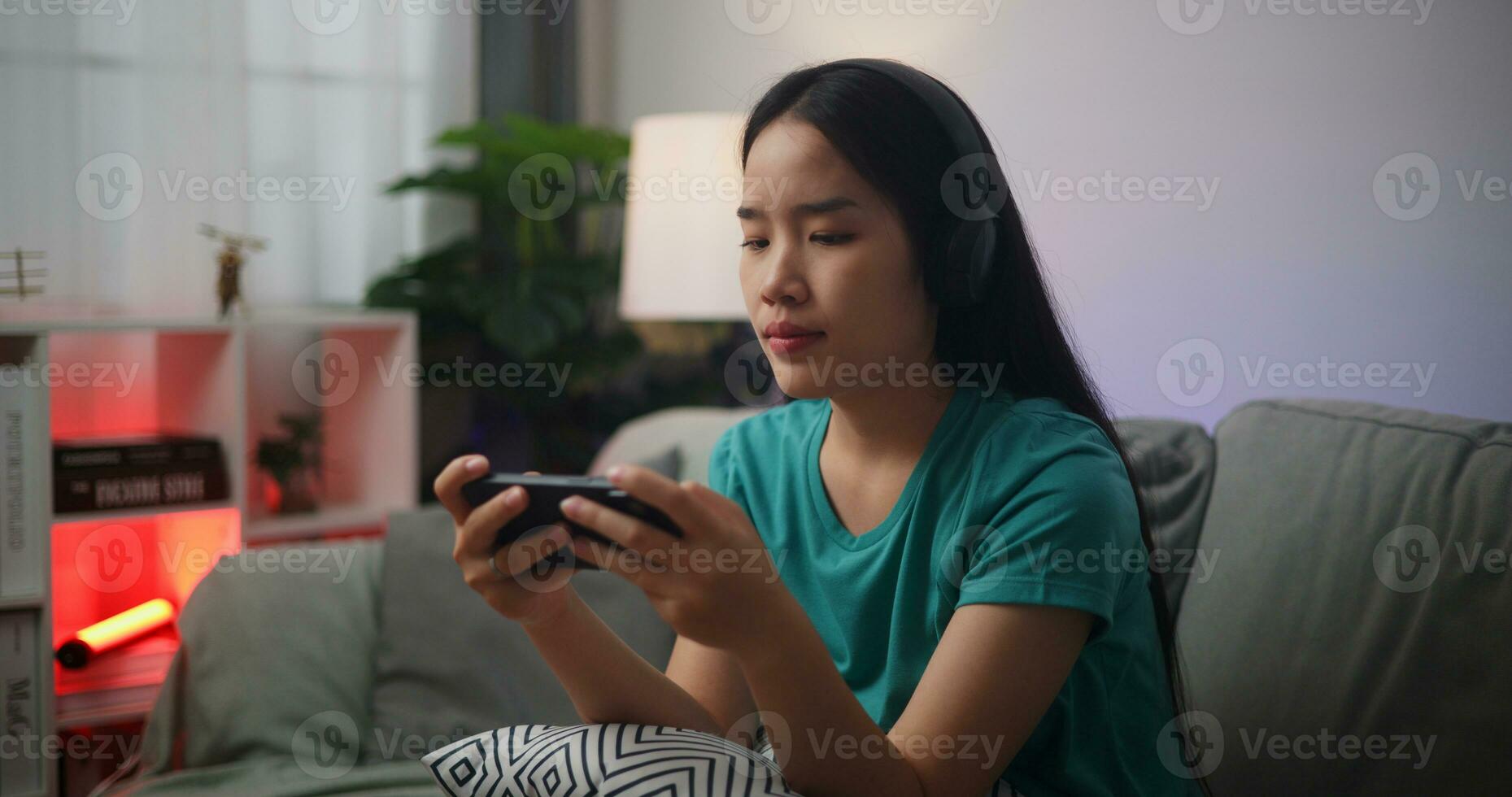 Portrait close up of Young Asian woman wearing glasses and headphones enjoys playing online esport games on smartphone sitting on sofa in the living room at home,Gamer lifestyle concept. photo