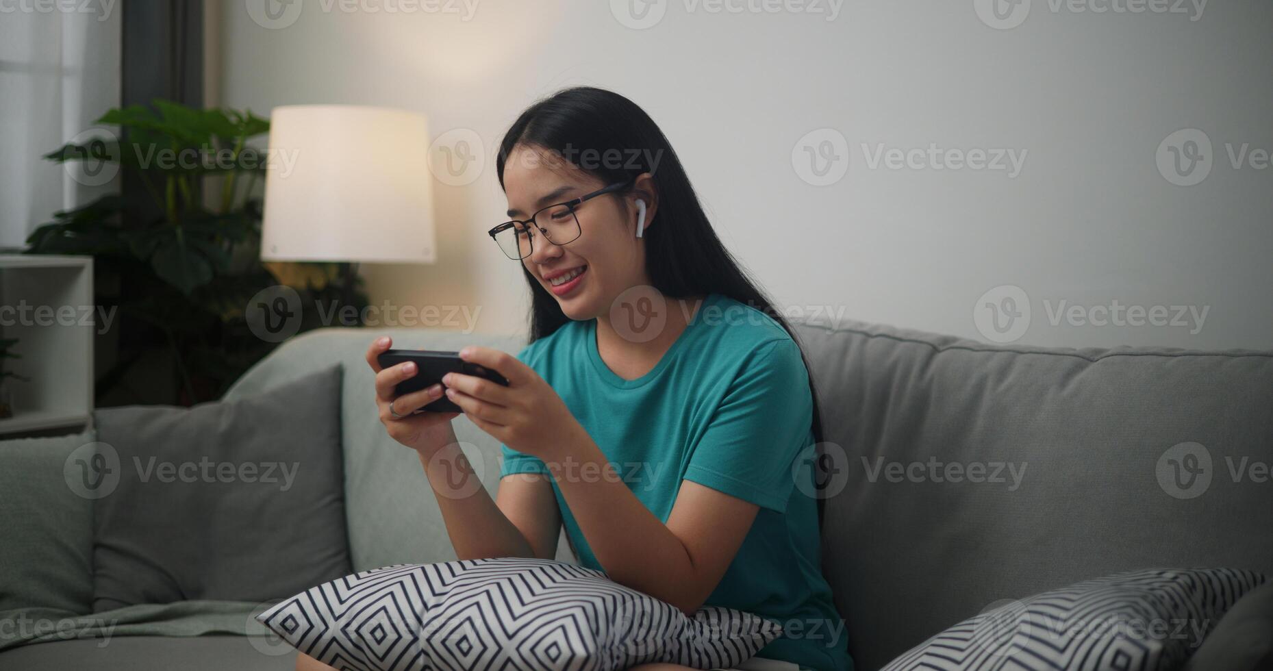 Portrait of Young Asian woman wearing glasses and headphones enjoys playing online esport games on smartphone sitting on sofa in the living room at home,Gamer lifestyle concept. photo
