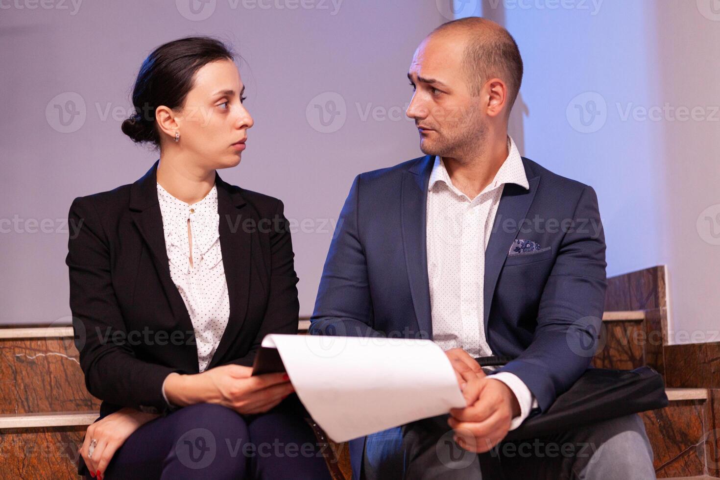 Employees on staircase late night afterhours tired from overtime, busy man and woman dealing with overwork and deadline. Colleague entrepreneur working late together in evening at corporate job sitting on staircase, explaining difficult finance project. photo