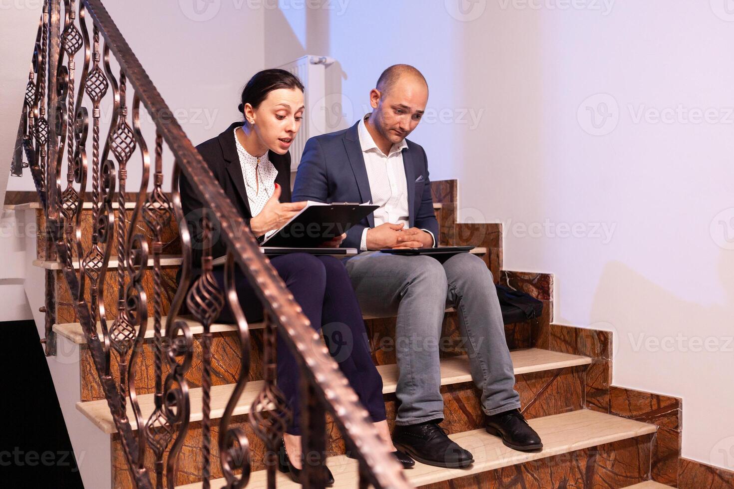 Worried tired businessman doing overtime at night on job diffuclt deadline with coworker sitting on stairs. Colleague entrepreneur working late together in evening at corporate job sitting on staircase, explaining difficult finance project. photo