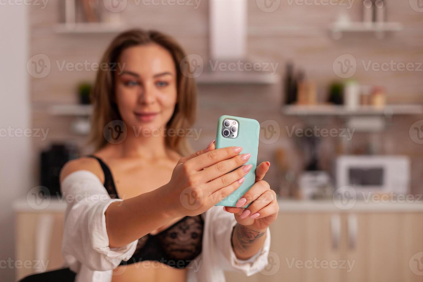 Portrait of beautiful woman looking at phone camera while taking selfie in home kitchen weraring sexy underwear. Seductive woman with tattoos using smartphone wearing temping lingerie in the morning. photo