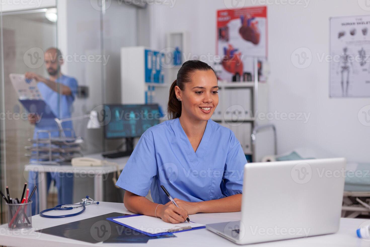 Smiling nurse using laptop computer and writing notes on clipboard in hospital office with doctor in the background. Health care physician using computer in modern clinic looking at monitor, photo