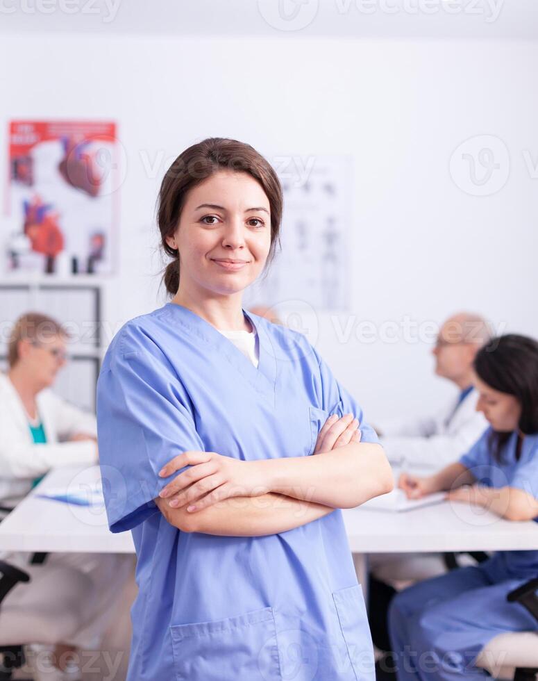 Medical therapist looking at camera with healthcare staff in the background wearing uniform. Friendly medical practitioner in clinic meeting room, robe, specialist. photo