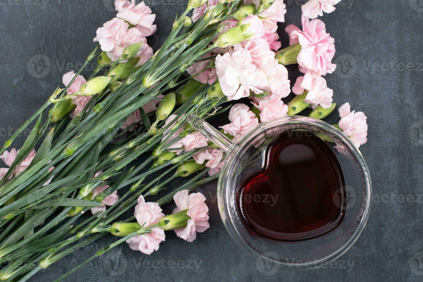 pink carnations on a dark background with a cup of red tea in a heart-shaped cup photo