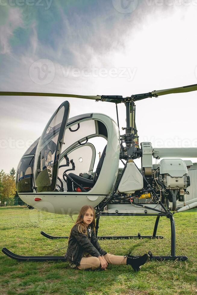 interpolación niña sentado en verde césped de volador campo cerca helicóptero foto