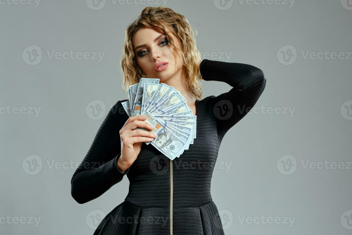 rubia doncella con Rizado cabello, en negro vestir es participación algunos dinero en efectivo, posando en gris antecedentes. juego entretenimiento, póker, casino. de cerca foto