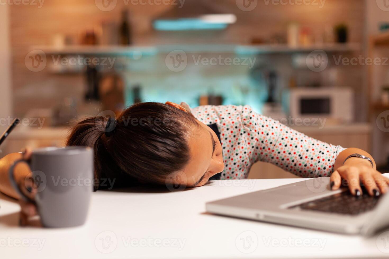 Freelance woman falling asleep in home kitchen working overtime on a project. Employee using modern technology at midnight doing overtime for job, business, busy, career, network, lifestyle ,wireless. photo