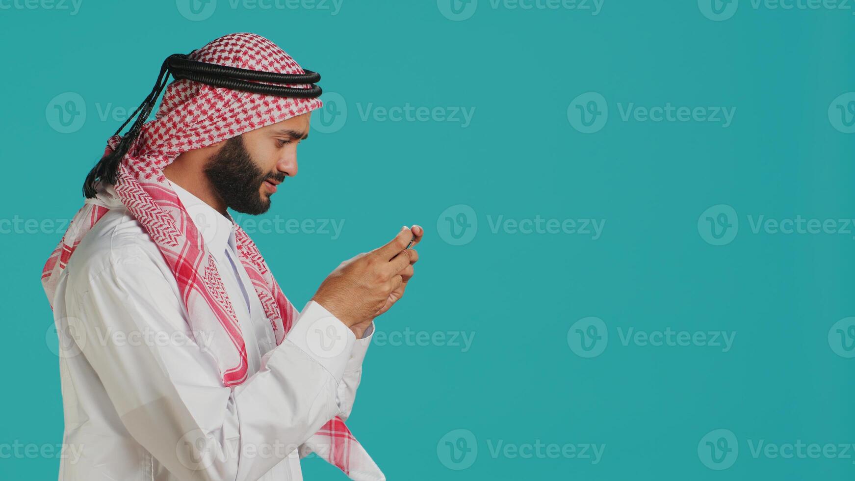 Joyful muslim man having fun with online game on his smartphone, wearing traditional islamic costume. In the studio, arab adult enjoying e sport challenge with mobile phone device. photo