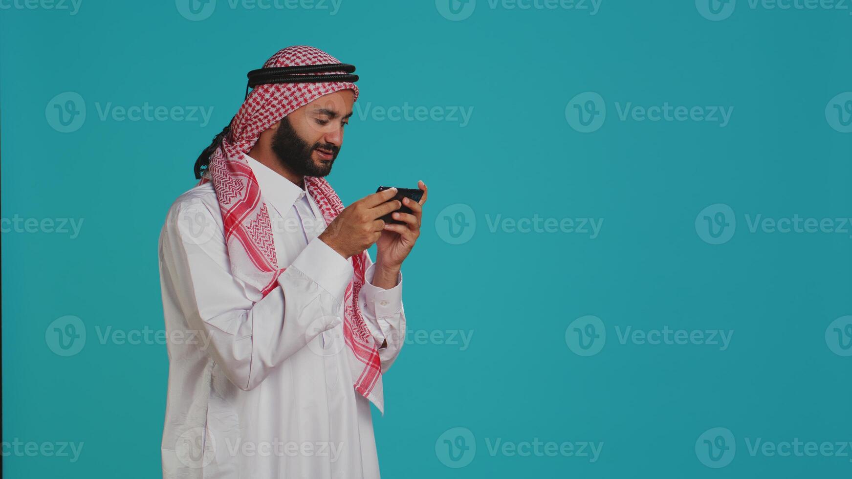Islamic guy playing fun mobile videogames on phone, dressed in traditional gown with headscarf. Middle eastern person engaging in online gaming contest using smartphone app. photo