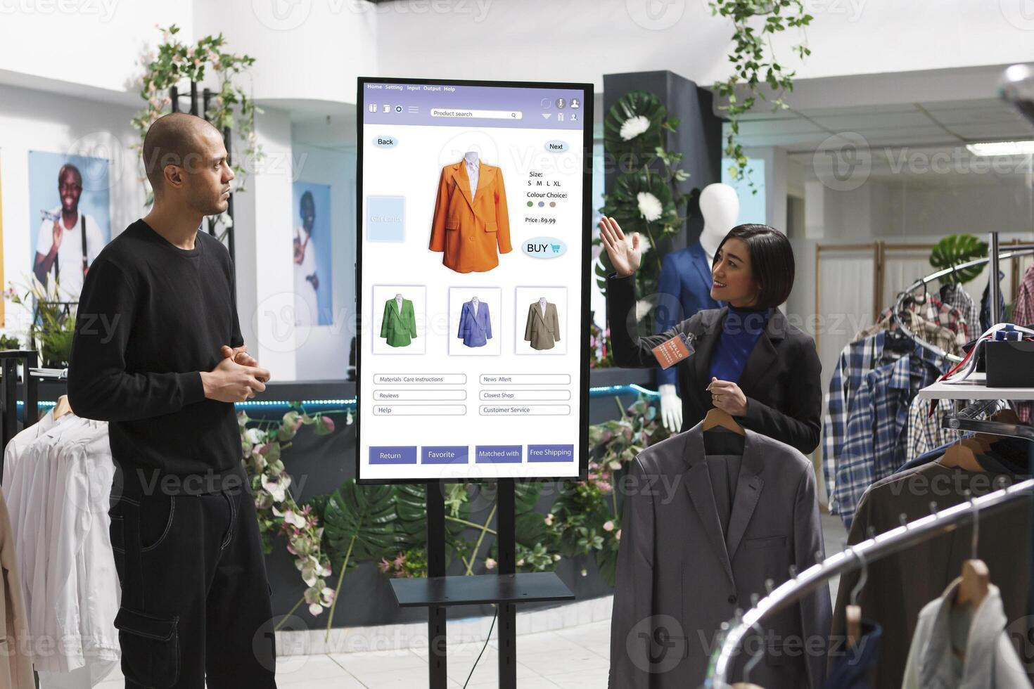Asian store assistant showcasing items to man in front of self service kiosk, selecting fashion collection clothes from clothing boutique. Caucasian customer discussing products with female employee. photo