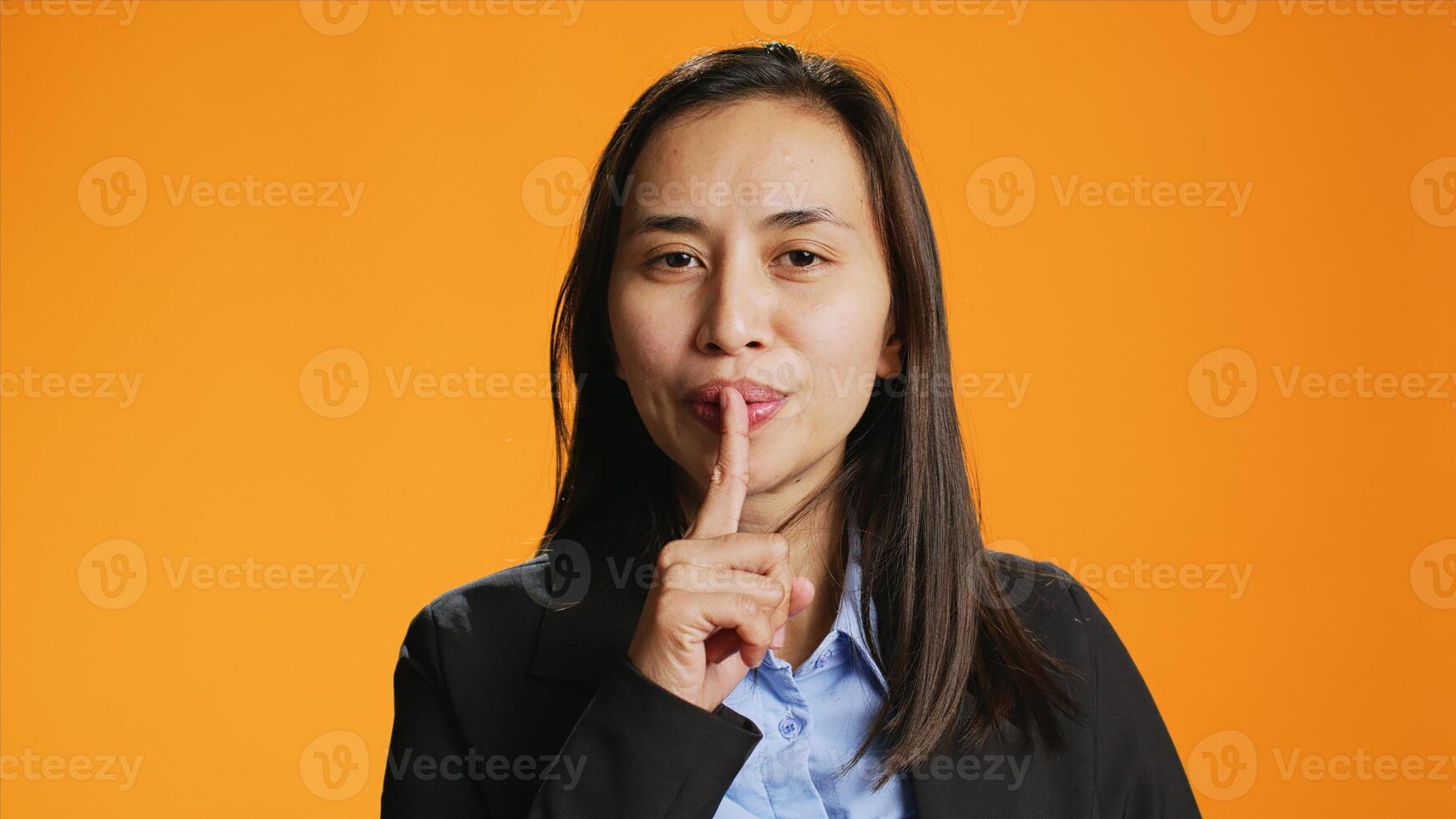Filipino person shows hush gesture on camera, doing mute sign to keep secret and be private. Woman putting finger over lips for silence, asking for secrecy against orange background. photo