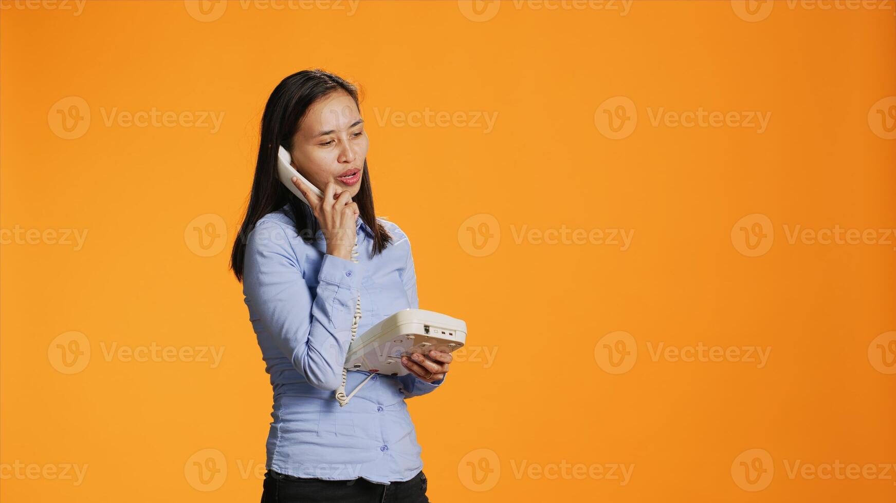 Asian woman using landline phone for remote call, talking to someone on vintage telephone line. Confident person answering office phone with cord, communicating remotely in studio. photo