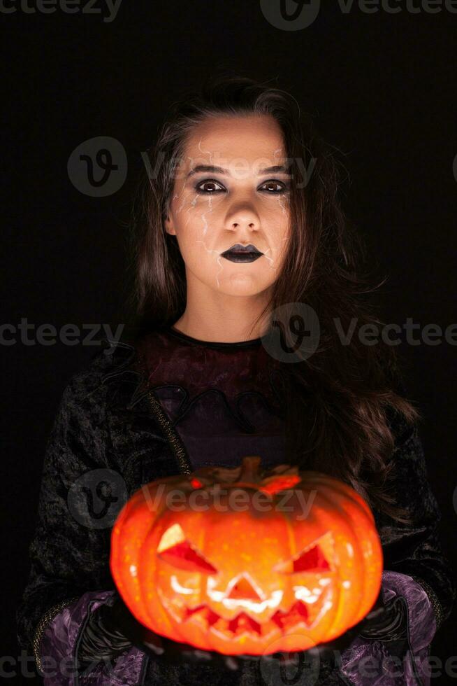 Attractive woman with a serious face dressed up like a witch holding a pumpkin over black background. photo