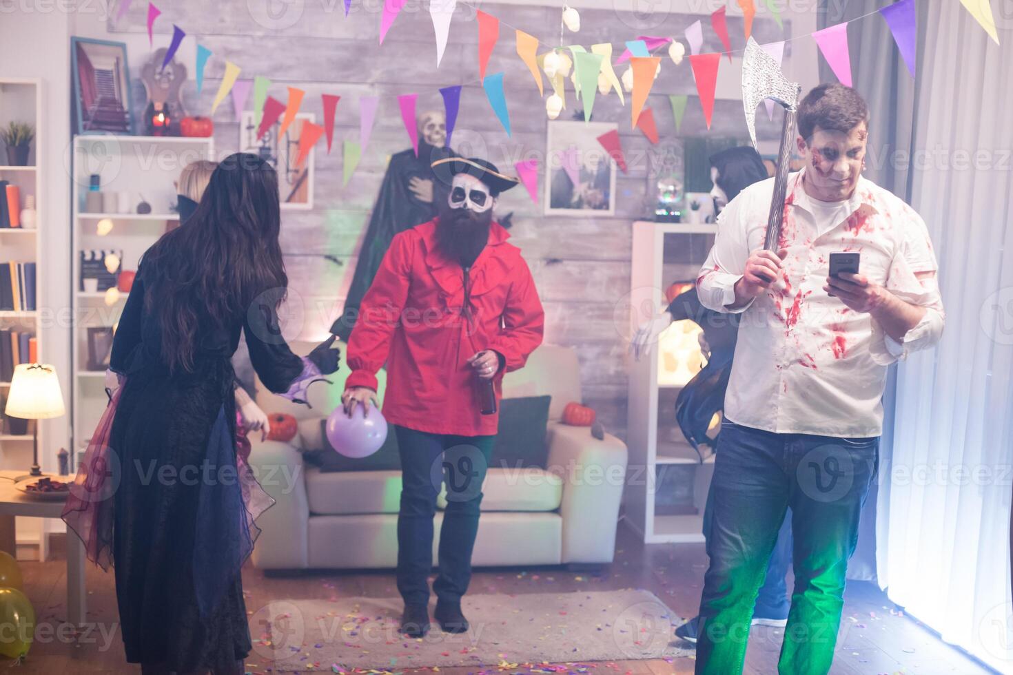 Young man dressed up like a zombie with an axe scrolling on his phone at halloween party. photo