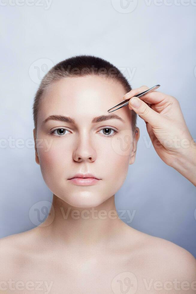 Young woman with short hair plucking eyebrows tweezers close up photo