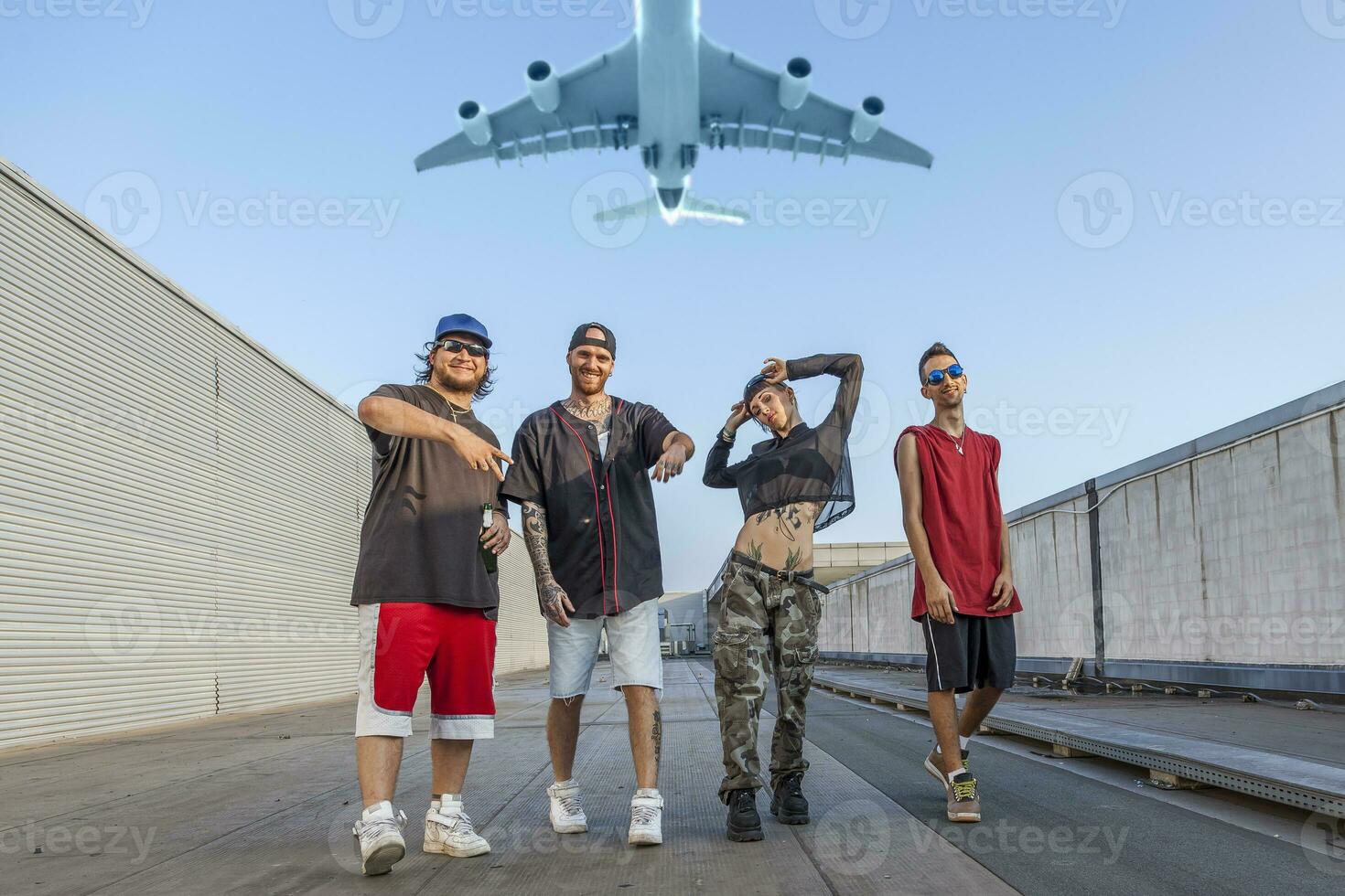 group of rappers posing on the metal rooftops of an abandoned building while a plane flies overhead photo