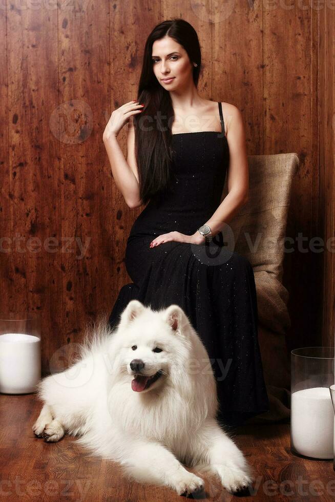 hermosa morena joven mujer largo negro vestir con un blanco como la nieve perro Samoyedo fornido estudio en sombras de marrón velas foto