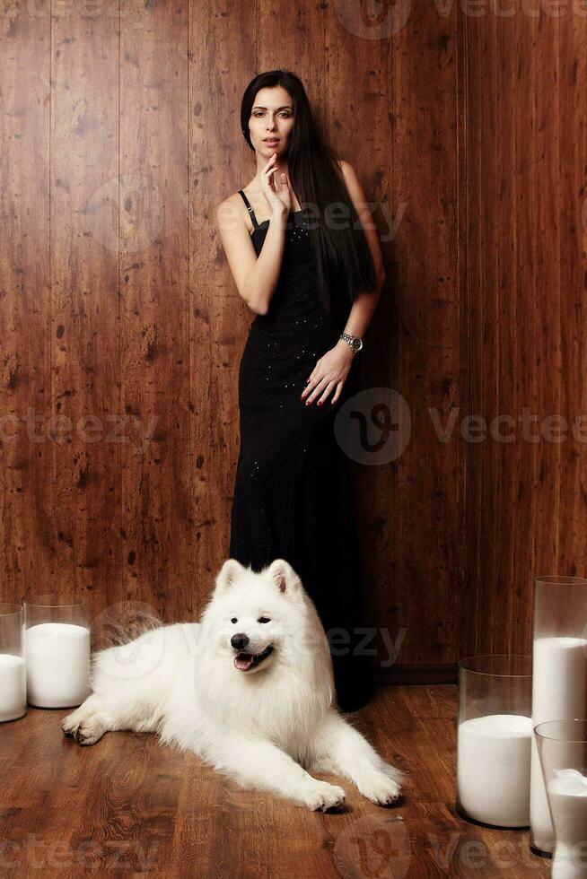 hermosa morena joven mujer largo negro vestir con un blanco como la nieve perro Samoyedo fornido estudio en sombras de marrón velas foto